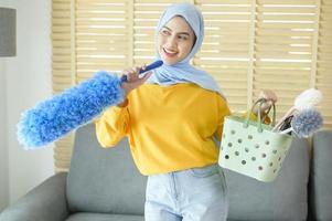 jonge gelukkige moslimvrouw die gele handschoenen draagt en een mand met schoonmaakmiddelen in de woonkamer vasthoudt. foto