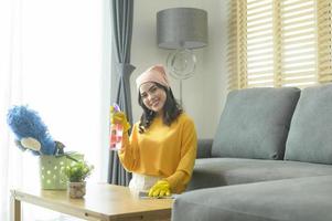 jonge gelukkige vrouw die gele handschoenen draagt en de tafel in de woonkamer afstoffen. foto