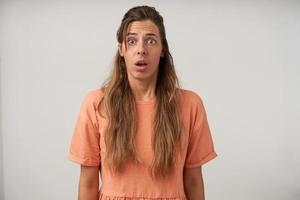 indoor shot van verbaasde jonge langharige vrouw die over een witte achtergrond staat met handen naar beneden, perzik t-shirt en casual make-up draagt, rimpelend met verrast gezicht foto