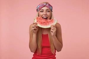 indoor shot van vrolijke mooie vrouw met hoofdband en lang blond haar poseren over roze achtergrond, watermeloen eten, in een goed humeur zijn foto