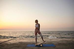 jonge sportieve blonde vrouw met lichaam in goede fysieke conditie poserend over zeezicht, sportieve kleding dragend, staande op balance board tijdens zonsopgang foto