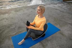 Jonge aantrekkelijke sportieve vrouw met blond haar die sporten aan zee maakt, op het balansbord zit en shaker met eiwit vasthoudt, bedachtzaam opzij kijkend foto