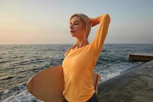 actieve jonge vrouw met blond haar poseren over zeezicht op de vroege ochtend, oranje top met lange mouwen dragen, houten plank vasthouden en hand op haar achterhoofd houden foto