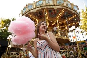 Positieve aantrekkelijke vrouw met lang bruin haar in zomerjurk poseren over carrousel in pretpark op warme zomerdag, suikerspin op houten stok houden, opzij kijkend en vrolijk glimlachen foto