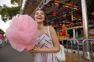 vrolijke jonge aantrekkelijke brunette dame met lang haar die door een pretpark loopt, zomerjurk draagt en suikerspin op stok houdt, opzij kijkt en vrolijk lacht foto