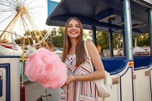 Positieve jonge aantrekkelijke brunette vrouw met lang haar poseren over pretpark op warme zonnige dag, romantische jurk en rugzak dragen, in de hand houden stok met roze suikerspin foto