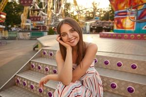 buitenportret van mooie jonge brunette vrouw in positieve stemming die haar hoofd met palm vasthoudt, zittend op een trap over park met attracties, lichtjes glimlachend naar de camera foto