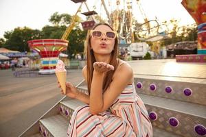 portret van aantrekkelijke jonge brunette vrouw met lang haar poseren buiten op warme zonnige dag, zittend op de trap en ijs in kegel houden, palm optillen en luchtkus blazen naar de camera foto