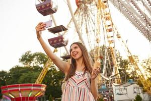 mooie langharige positieve vrouw die over het reuzenrad staat met een ijsje terwijl ze selfie maakt op haar mobiele telefoon, in een goed humeur is en vrolijk lacht foto