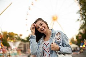 jonge mooie vrolijke brunette vrouw in trendy jeans jas staande over reuzenrad in pretpark, gelukkig naar de camera kijkend en haar haar rechttrekken foto