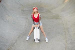 buitenfoto van mooie langharige getatoeëerde blonde vrouw poserend over skatepark, leunend aan boord en ogen gesloten houdend, casual kleding dragend foto