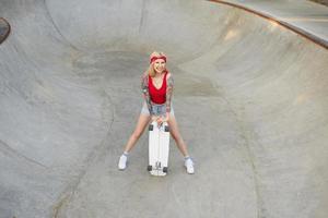 vrolijke langharige getatoeëerde blonde vrouw die zich voordeed boven skatepark op warme heldere dag, jeans shorts en rode top dragen, bord in handen houden en gelukkig glimlachen foto