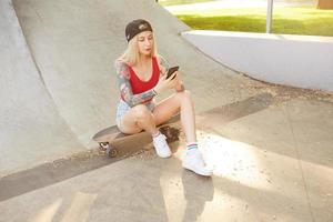 Zonnig buitenschot van aantrekkelijke jonge blonde vrouw met tatoeages zittend boven stadspark op skateboard, rode top en spijkerbroek dragend, smartphone in handen houdend foto