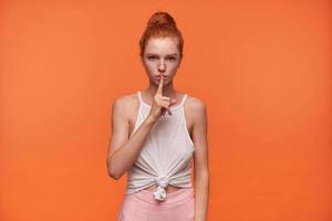 studioportret van mooie jonge vrouw die haar rode haar in een knoop draagt, wijsvinger op haar lippen houdt, vraagt om geheim te houden, geïsoleerd over oranje achtergrond in vrijetijdskleding foto