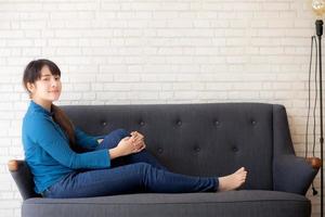 mooi portret jonge aziatische vrouw zitten en glimlachen gelukkig en kijken naar de camera op de bank met casual in de woonkamer, meisje vrolijk en ontspannen op de bank thuis, lifestyle concept. foto