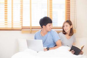 Aziatisch jong stel met man met laptopcomputer en zoeken op internet en vrouw die notitieboekje leest op bed in de slaapkamer met gelukkig en ontspannen, communicatie en bedrijfsconcept. foto