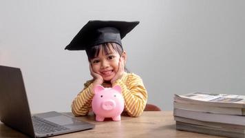 Aziatisch klein meisje met een afstudeerpet met een roze spaarvarken, geld besparen, investeren in de toekomst, foto