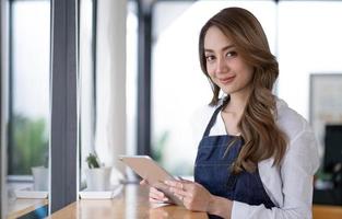 startup succesvolle kleine ondernemer mkb schoonheid meisje staan met tablet smartphone in coffeeshop restaurant. portret van aziatische tan vrouw barista café-eigenaar. MKB ondernemer verkoper bedrijfsconcept foto