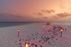 bestemming dineren, huwelijksreis, jubileum. geweldig romantisch diner strand op houten dek met kaarsen onder avondrood. romantiek en liefde, luxe dineren op een bestemming, exotische tafelopstelling met uitzicht op zee foto