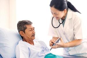 Aziatische vrouw professionele arts met klembord bezoeken, praten en diagnosticeren van de oude man patiënt liggend in patiëntenbed op ziekenhuisafdeling foto