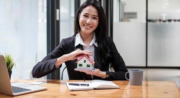 miniatuur huis in de handen van een aziatische vrouw makelaar woningkrediet werken op kantoor. naar de camera kijken. foto