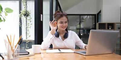 gelukkig jong aziatisch tienermeisje zit thuis op de bank met telefoon en kijkt naar het scherm zwaaiende hand videobellen op afstand vriend online in laptop met behulp van laptop videochat-applicatie. foto