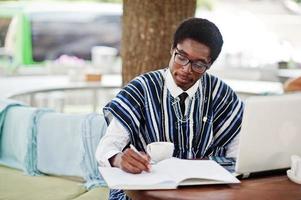 afrikaanse man in traditionele kleding en bril zit aan de buitencaffe, drinkt koffie achter de laptop en schrijft iets op zijn notitieboekje. foto