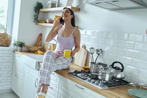 gelukkige vrouw praten op mobiele telefoon en genieten van koffie zittend op het aanrecht foto