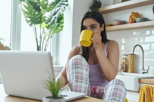 aantrekkelijke jonge vrouw die van koffie in de ochtend geniet en laptop gebruikt bij de binnenlandse keuken foto