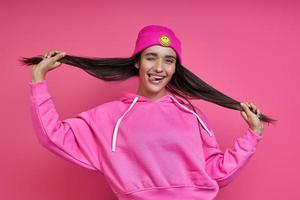 speelse jonge vrouw in overhemd met capuchon die haar lange haar tegen roze achtergrond houdt foto