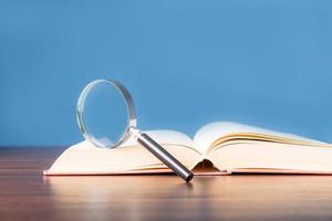 open boek met vergrootglas op houten bureau in informatiebibliotheek van school of universiteit, concept voor onderwijs, lezen, studeren, kopiëren van ruimte en blauwe achtergrond. foto