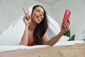 speelse jonge vrouw die zich bedekt met een deken terwijl ze selfie maakt met een smartphone in bed foto