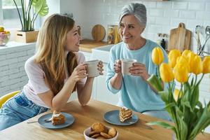 senior moeder en haar volwassen dochter genieten van warme dranken en zoet eten in de keuken foto