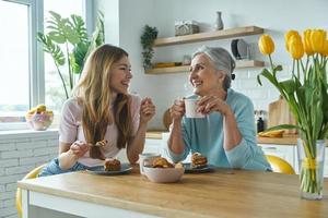 senior moeder en haar volwassen dochter genieten van warme dranken en zoet eten in de keuken foto