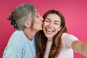 senior moeder kust haar vrolijke dochter terwijl ze selfie maakt tegen roze achtergrond foto