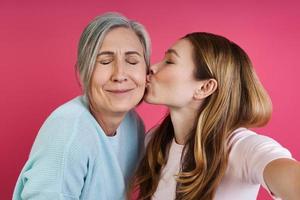 volwassen dochter kust haar oudere moeder terwijl ze selfie maakt tegen roze achtergrond foto