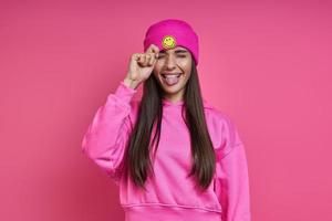 speelse vrouw in overhemd met capuchon die haar funky hoed aanpast en een gezicht trekt tegen een roze achtergrond foto