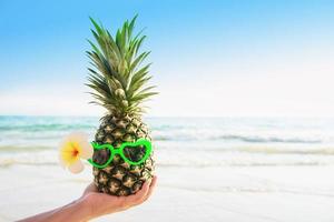 heerlijke verse ananas die een bril in toeristische handen zet met zeegolfachtergrond - gelukkig plezier met gezond vakantieconcept foto