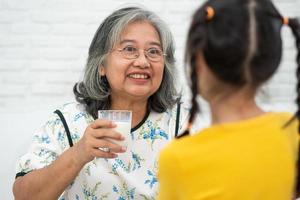 gelukkige bejaarde aziatische oma zit naast haar kleindochter en voedt thuis verse melk uit glas voor het ontbijt. concept van een gelukkig gezin en zorgt samen, voorschoolse gezondheidszorg foto