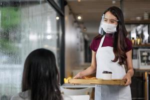 aziatische serveerster met gezichtsbeschermend masker en gezichtsschild dat koffie en brood serveert in de coffeeshop. foto