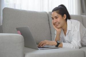 vrolijke lachende aziatische vrouw die laptop gebruikt terwijl ze op de bank in de woonkamer zit. ze gebruikt haar laptop om te vergaderen, te zoeken, te studeren en te chatten met vrienden en om online te winkelen. foto