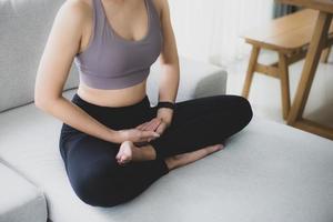 vrouw met zwarte sportkleding die op de bank zit en yoga beoefent en mediteert in de woonkamer. ze bereidt materiaal voor voor de oefenles. rust en ontspanning. vrouwelijk geluk en yogaconcept. foto