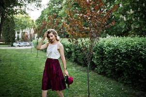 modieuze en mooie blonde model meisje in stijlvolle rood fluwelen velours rok, witte blouse en hoed, geposeerd in het park met telefoon en koptelefoon. foto