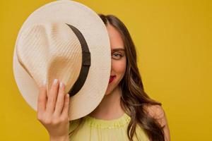 close-up portret van een vrolijke bruinharige vrouw met lichte make-up hoed houden op haar gezicht geïsoleerd op gele achtergrond. ware emoties, geluk foto