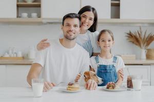 gelukkige familie en hond poseren in een gezellige keuken, eet verse zelfgemaakte pannenkoeken met chocolade en melk, kijk positief naar de camera. moeder in schort omhelst man en dochter, kookt graag voor hen foto