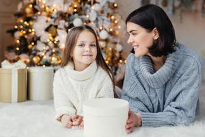 winter en feest concept. mooie mooie blauwe ogen klein vrouwelijk kind in gebreide witte trui en brunette vrouw hebben goede rust thuis, tijd doorbrengen in de buurt van versierde kerstboom foto