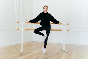 horizontaal schot van flexibele jonge vrouw leunt op ballet barre, draagt een zwart kostuum en witte sportschoenen, glimlacht positief, poseert in de danszaal. balletleraar demonstreert verschillende oefeningen foto