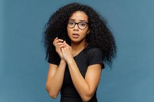 foto van beschaamde afro-amerikaanse vrouw houdt handen bij elkaar, kijkt nerveus naar de camera, heeft wat problemen, draagt een casual zwart t-shirt, staat tegen een blauwe achtergrond. negatieve gevoelens concept