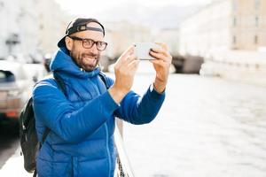 stijlvolle man met blauwe charmante ogen en baard met blauwe anorak, brillen en pet die selfie maken met mobiele telefoon tegen de achtergrond van de grote stad. jonge toerist met rugzak die foto van zichzelf maakt