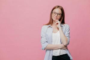 indoor shot van bedachtzame blanke vrouw houdt één hand onder de kin, gekleed in een elegant shirt, modellen over roze achtergrond, geïsoleerd over roze achtergrond met kopie ruimte voor uw promotie of slogan foto